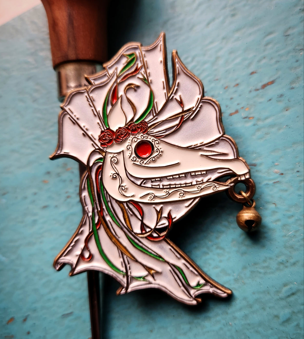 Mari Lwyd lapel pin - horse skull holding bell against white sheet with red, green, and gold ribbons